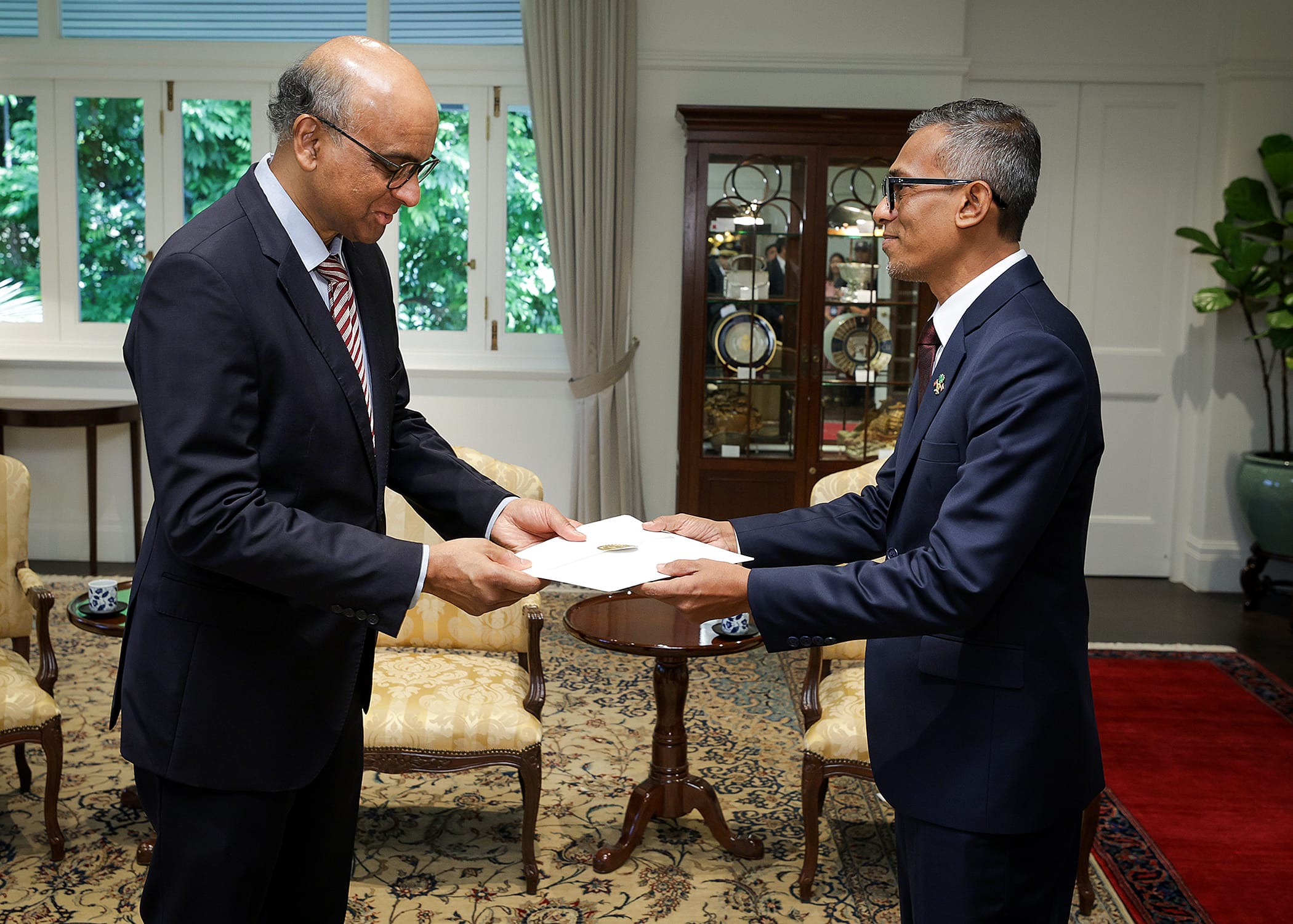 Two men in suits exchanging a document in an ornate room with a red rug.